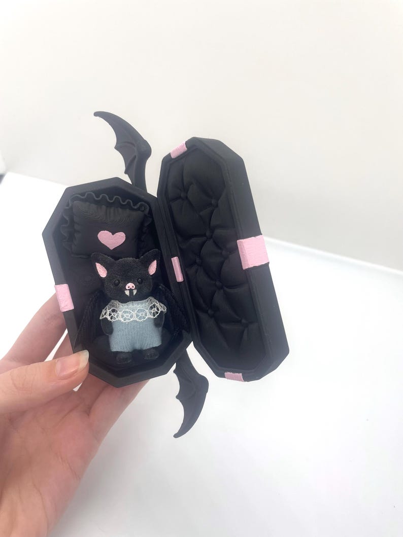 以下が含まれることがあります： ピンクのアクセントが付いた黒い棺型の箱で、開くとぬいぐるみコウモリの人形が現れます。 コウモリは青い衣装とレースの襟を着ており、ピンクのハートが付いた枕の上にいます。 棺にはコウモリの羽のディテールがあります。