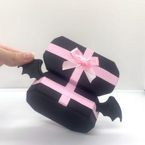 以下が含まれることがあります： ピンクのリボンとリボンが付いた、2つの積み重ねられた黒いギフトボックス。各ボックスには、側面に黒いコウモリの羽が1組付いています。ボックスは白い表面にあります。