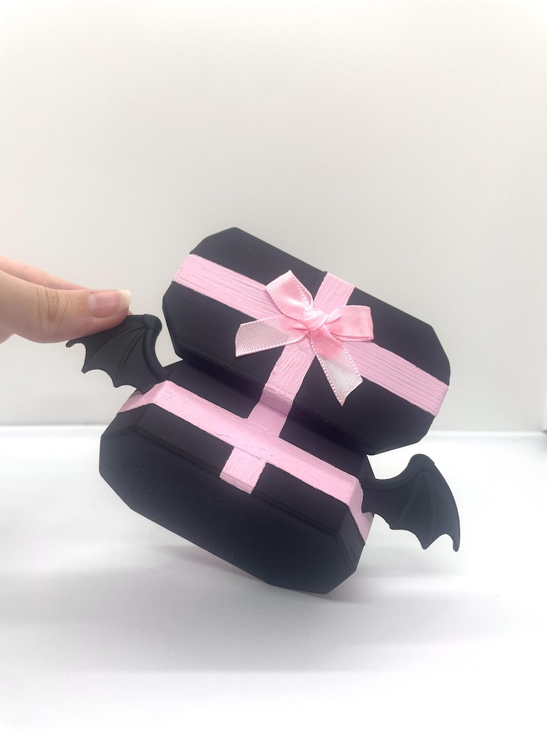 以下が含まれることがあります： ピンクのリボンとリボンが付いた、2つの積み重ねられた黒いギフトボックス。各ボックスには、側面に黒いコウモリの羽が1組付いています。ボックスは白い表面にあります。