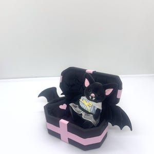 以下が含まれることがあります： ピンクの耳とドレスを着た黒いぬいぐるみコウモリが、ピンクのディテールが施された黒い棺の中に座っています。 棺にはピンクのハートとコウモリの羽が付いています。 このゴシックをテーマにしたアイテムは、ユニークな装飾品です。