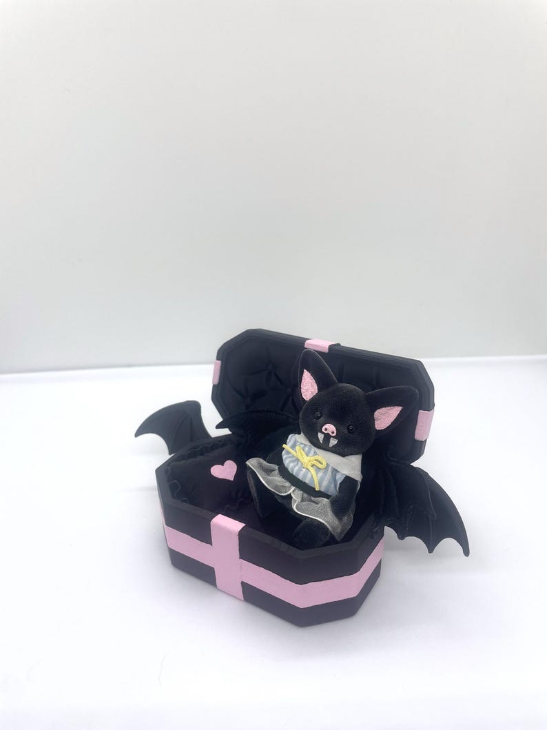以下が含まれることがあります： ピンクの耳とドレスを着た黒いぬいぐるみコウモリが、ピンクのディテールが施された黒い棺の中に座っています。 棺にはピンクのハートとコウモリの羽が付いています。 このゴシックをテーマにしたアイテムは、ユニークな装飾品です。