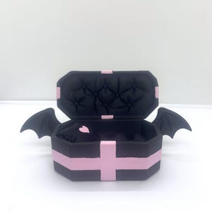 以下が含まれることがあります： ピンクのアクセントとコウモリの羽が付いた、黒い棺型の箱。内側はキルティングデザインの黒色です。開いた箱の中にピンクのハートが見えます。箱は白い表面にあります。