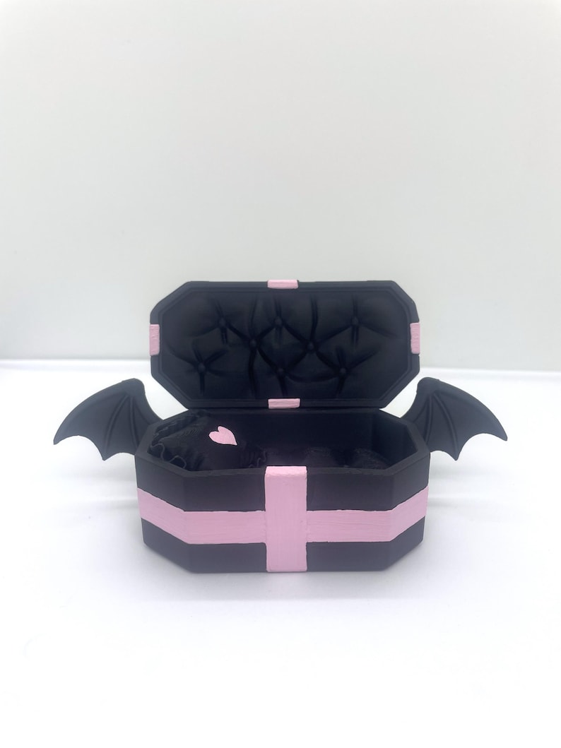 以下が含まれることがあります： ピンクのアクセントとコウモリの羽が付いた、黒い棺型の箱。内側はキルティングデザインの黒色です。開いた箱の中にピンクのハートが見えます。箱は白い表面にあります。
