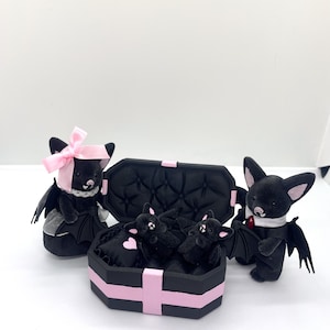 以下が含まれることがあります： ピンクのアクセントが付いた黒いぬいぐるみコウモリのコレクション。ピンクのリボンをつけたコウモリ、タキシードを着たコウモリ、棺型の箱に入ったコウモリなど。箱は黒とピンクのトリムです。
