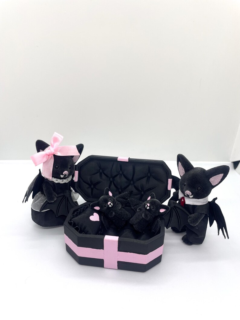 以下が含まれることがあります： ピンクのアクセントが付いた黒いぬいぐるみコウモリのコレクション。ピンクのリボンをつけたコウモリ、タキシードを着たコウモリ、棺型の箱に入ったコウモリなど。箱は黒とピンクのトリムです。