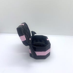 以下が含まれることがあります： ピンクのストライプとキルティング加工の内装が施された、開いた黒いジュエリーボックス。ボックスは八角形で、側面に黒いリボンが付いています。内側は黒で、リングホルダーが付いています。