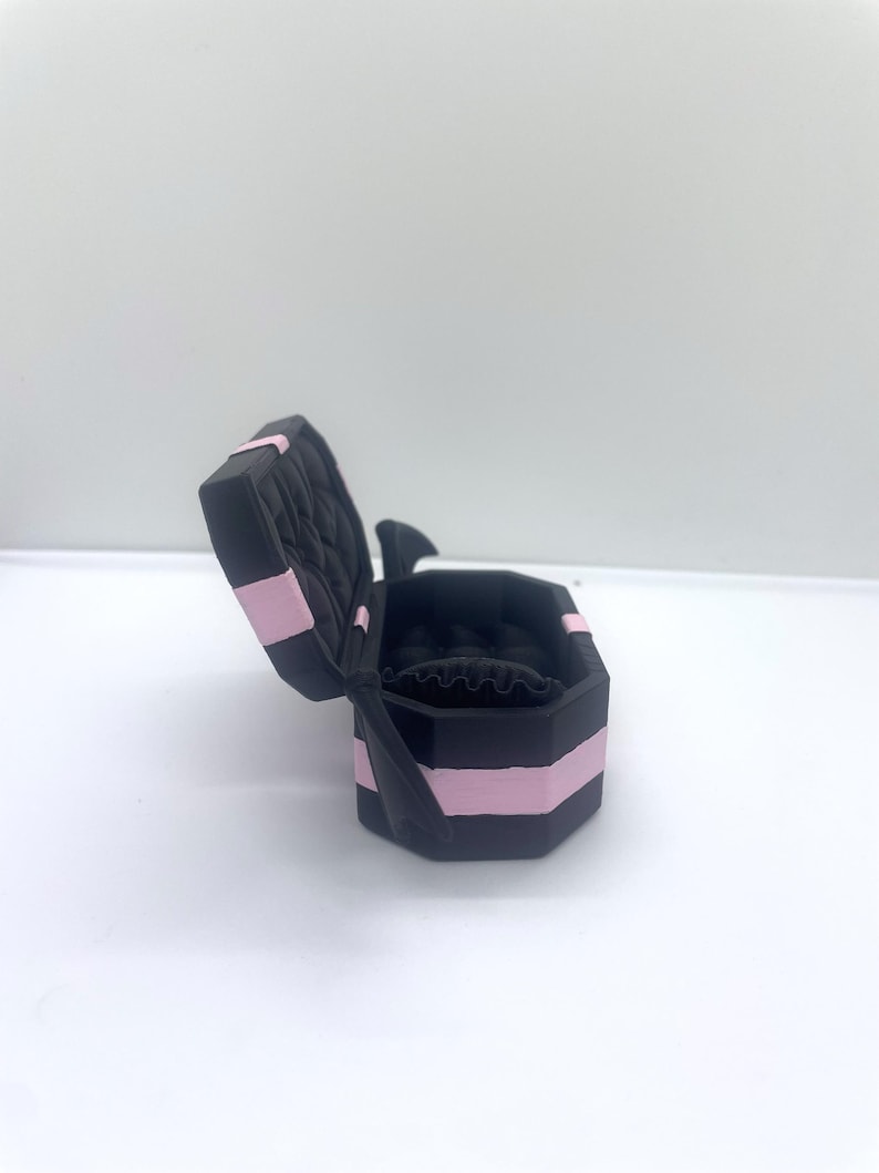 以下が含まれることがあります： ピンクのストライプとキルティング加工の内装が施された、開いた黒いジュエリーボックス。ボックスは八角形で、側面に黒いリボンが付いています。内側は黒で、リングホルダーが付いています。