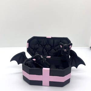 以下が含まれることがあります： ピンクのアクセントとキルティング加工の内装が施された黒い棺型の箱。羽を広げた黒いぬいぐるみコウモリが複数入っています。棺は開いており、コウモリが見えています。