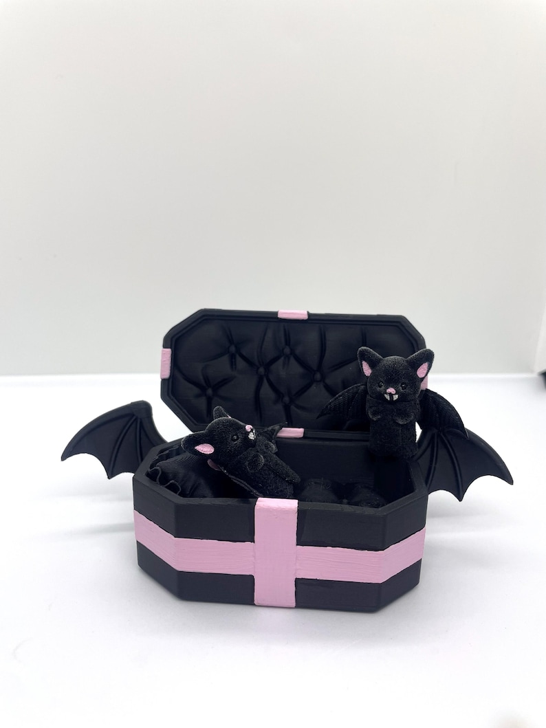 以下が含まれることがあります： ピンクのアクセントとキルティング加工の内装が施された黒い棺型の箱。羽を広げた黒いぬいぐるみコウモリが複数入っています。棺は開いており、コウモリが見えています。