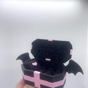 以下が含まれることがあります： ピンクのアクセントとコウモリの羽が付いた、黒い棺型の箱。箱は開いており、内側にハート型のディテールが現れています。棺はピンクのストライプとピンクの十字架で飾られています。