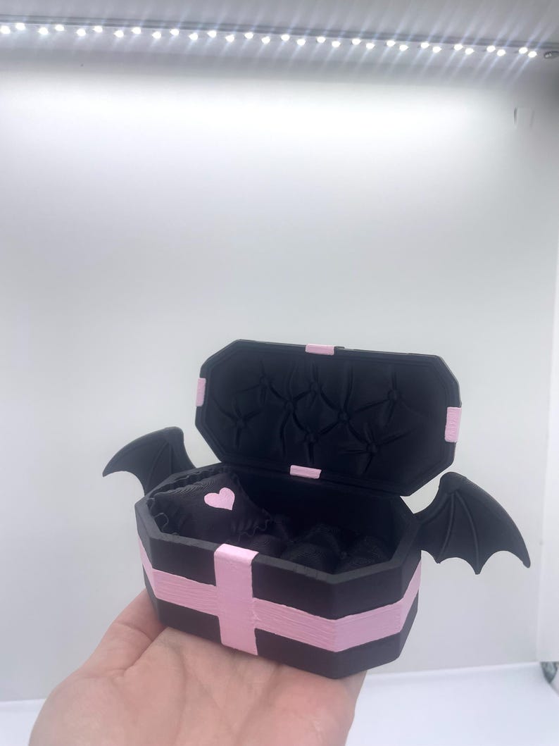 以下が含まれることがあります： ピンクのアクセントとコウモリの羽が付いた、黒い棺型の箱。箱は開いており、内側にハート型のディテールが現れています。棺はピンクのストライプとピンクの十字架で飾られています。