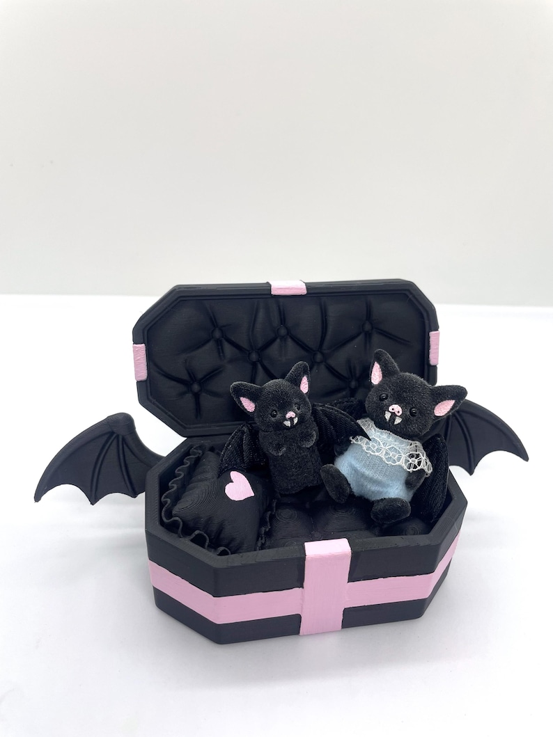 以下が含まれることがあります： ピンクのアクセントとコウモリの羽が付いた黒い棺型の箱で、2匹のぬいぐるみコウモリが入っています。コウモリの1匹はレーストリムの青いドレスを着ています。棺の内側は黒色のタフティングです。ピンクのハートが付いた小さな枕も入っています。