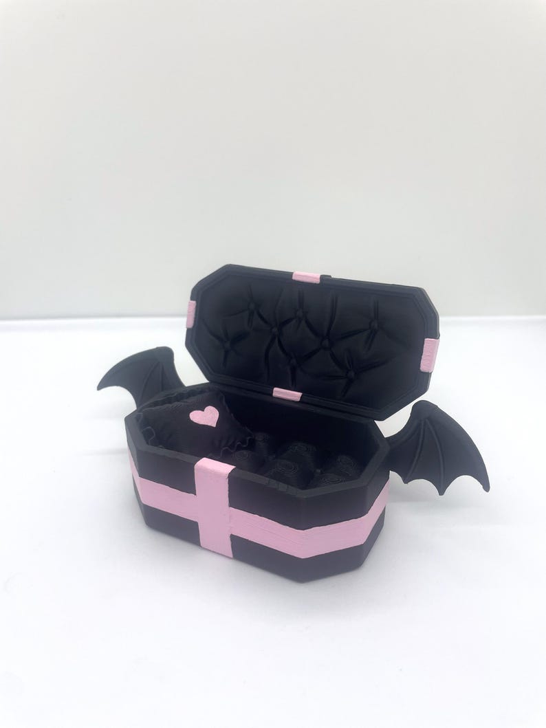 以下が含まれることがあります： ピンクのアクセントとコウモリの羽が付いた、黒い棺型の箱。箱は開いており、ピンクのハートが付いた黒い内側が見えます。棺はピンクのストライプとアクセントで装飾されています。