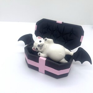 以下が含まれることがあります： ピンクのアクセントとコウモリの羽が付いた黒い棺の中に、白いブルドッグの置物が横たわっています。棺の内側はタフテッド加工が施されています。置物には黒い足パッドが付いています。