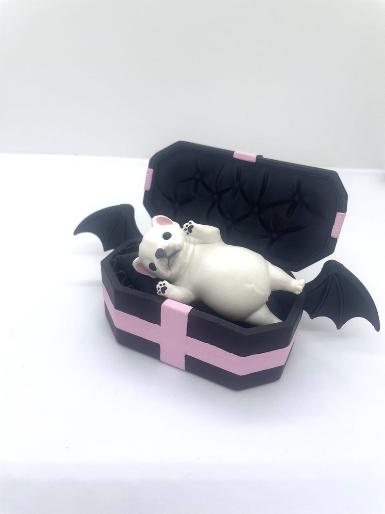 以下が含まれることがあります： ピンクのアクセントとコウモリの羽が付いた黒い棺の中に、白いブルドッグの置物が横たわっています。棺の内側はタフテッド加工が施されています。置物には黒い足パッドが付いています。