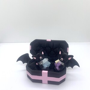 以下が含まれることがあります： ピンクのアクセントが付いた黒い棺型の箱の中に、2つのぬいぐるみコウモリのフィギュアが入っています。コウモリは黒い毛皮、ピンクの耳を持ち、パステルカラーの衣装を着ています。棺には黒いコウモリの羽が付いています。