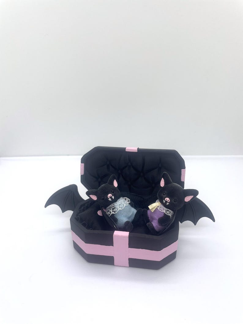 以下が含まれることがあります： ピンクのアクセントが付いた黒い棺型の箱の中に、2つのぬいぐるみコウモリのフィギュアが入っています。コウモリは黒い毛皮、ピンクの耳を持ち、パステルカラーの衣装を着ています。棺には黒いコウモリの羽が付いています。