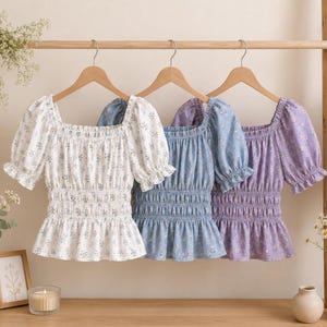 May include: Three floral print tops in white, blue, and purple hang on a wooden rod. Each top has a square neckline, short puff sleeves, and a shirred waist. The tops are decorated with small floral patterns.