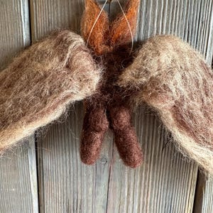 Può includere: Ornamento a forma di angelo fatto a mano in lana marrone e beige. L'angelo presenta ali spiegate, un corpo centrale e una testa con orecchie a punta. È sospeso da un filo sottile e un piccolo disco di legno.