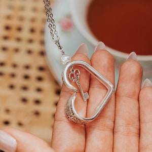 May include: A silver-toned heart-shaped pendant with a small key charm inside, suspended from a delicate chain. A faux pearl accents the chain. The pendant is held in a hand, with a teacup in the background.