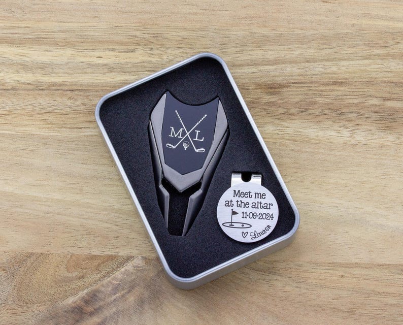 Golf ball marker and divot tool set in a gift tin box. Divot tool has marker engraved with crossed clubs with an initial on each side. The round marker comes with a hat clip and is engraved Meet me at the altar with the date, golf green graphic and a heart with name at the bottom.