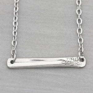 May include: A silver necklace featuring a delicate chain and a bar pendant with a floral design.