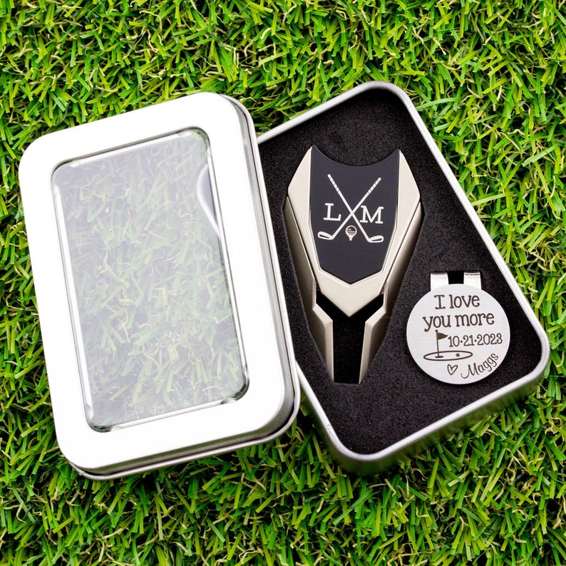 Golf ball marker and divot tool set in a gift tin box. Divot tool has marker engraved with crossed clubs with an initial on each side. The round marker comes with a hat clip and is engraved love you more with the date, golf green graphic and a heart with name at the bottom.