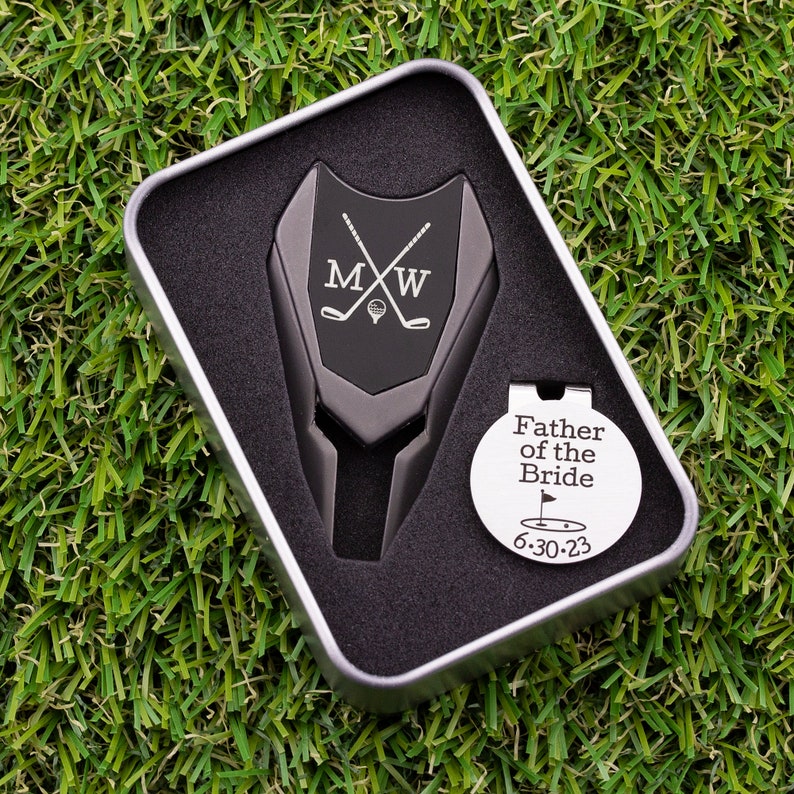 Golf ball marker and divot tool set in a gift tin box. Divot tool has marker engraved with crossed clubs with an initial on each side. The round marker comes with a hat clip and is engraved Father of the bride with the date and golf green graphic.
