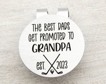 May include: Silver golf ball marker with the text "The Best Dads Get Promoted To Grandpa Est. 2023" and a pair of crossed golf clubs with a golf ball.
