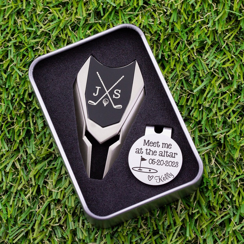 Golf ball marker and divot tool set in a gift tin box. Divot tool has marker engraved with crossed clubs with an initial on each side. The round marker comes with a hat clip and is engraved Meet me at the altar with the date, golf green graphic and a heart with name at the bottom.