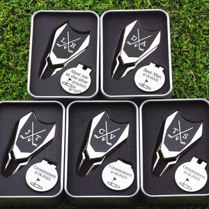 This image shows 5 Golf ball marker and divot tool sets in a gift tin boxes. Divot tool has marker engraved with crossed clubs with an initial on each side. The round marker comes with a hat clip and can be engraved Meet me at the altar with the date, golf green graphic and a heart with name at the bottom for the groom from bride, Groomsman with date and golf graphic, Best Man with date and golf graphic. Gifts for the wedding party.