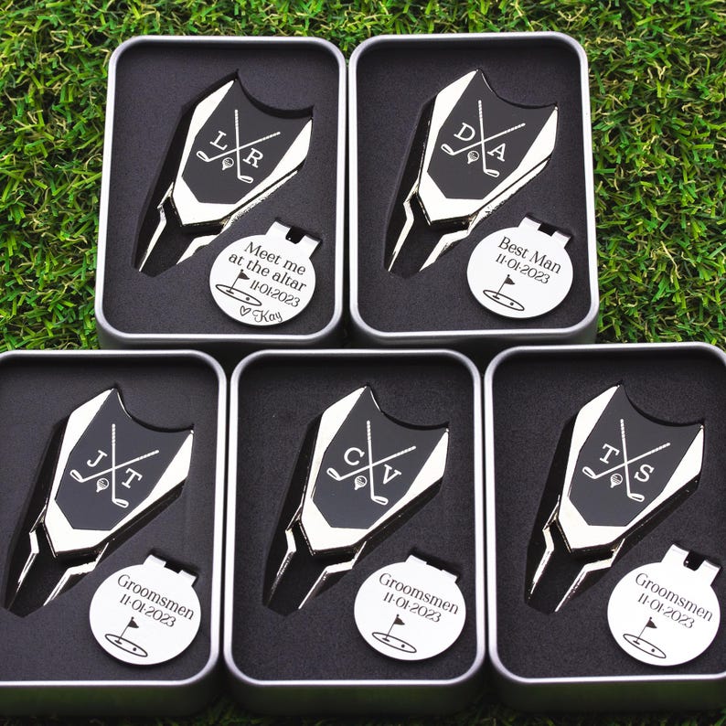 This image shows 5 Golf ball marker and divot tool sets in a gift tin boxes. Divot tool has marker engraved with crossed clubs with an initial on each side. The round marker comes with a hat clip and can be engraved Meet me at the altar with the date, golf green graphic and a heart with name at the bottom for the groom from bride, Groomsman with date and golf graphic, Best Man with date and golf graphic. Gifts for the wedding party.