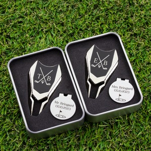 Golf ball marker and divot tool set in a gift tin box. Divot tool has marker engraved with crossed clubs with an initial on each side. The round marker comes with a hat clip and is engraved with Mr Name and Mrs Name with date and golf green graphic.