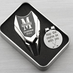 Golf ball marker and divot tool set in a gift tin box. Divot tool has marker engraved with a large M with a name through the center of it and Groomsman and the date under it. The round marker comes with a hat clip and is engraved may the course be with you and crossed clubs graphic.