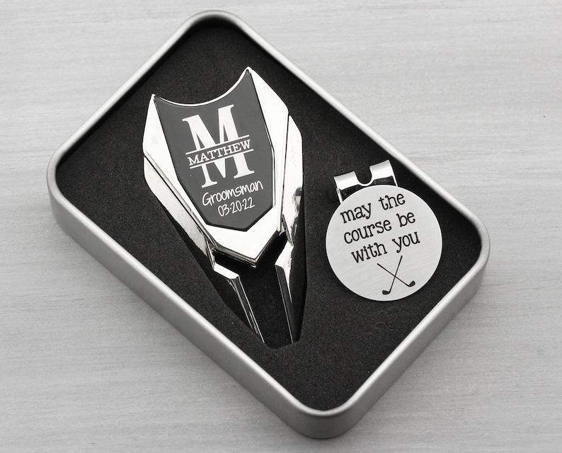 Golf ball marker and divot tool set in a gift tin box. Divot tool has marker engraved with a large M with a name through the center of it and Groomsman and the date under it. The round marker comes with a hat clip and is engraved may the course be with you and crossed clubs graphic.