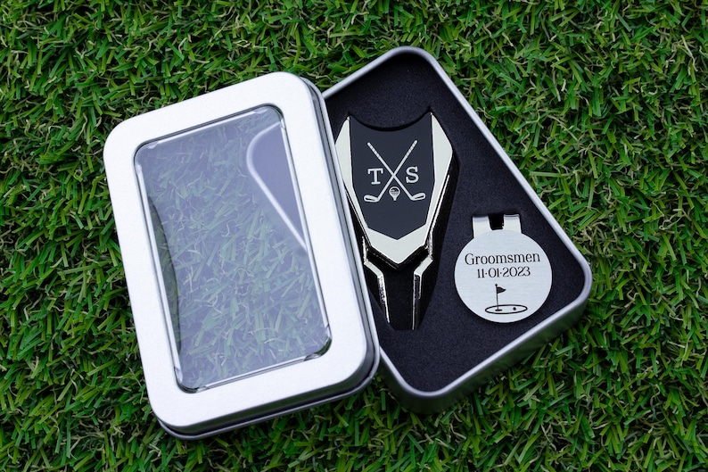 Golf ball marker and divot tool set in a gift tin box. Divot tool has marker engraved with crossed clubs with an initial on each side. The round marker comes with a hat clip and is engraved Groomsmen with the date and golf green graphic.