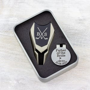 Golf ball marker and divot tool set in a gift tin box. Divot tool has marker engraved with crossed clubs with an initial on each side. The round marker comes with a hat clip and is engraved father of the bride with the date and golf green graphic.