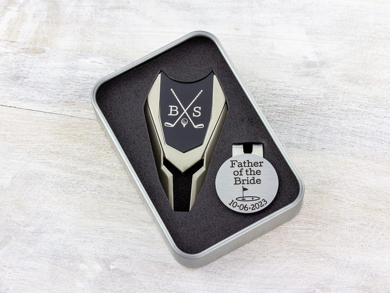 Golf ball marker and divot tool set in a gift tin box. Divot tool has marker engraved with crossed clubs with an initial on each side. The round marker comes with a hat clip and is engraved father of the bride with the date and golf green graphic.