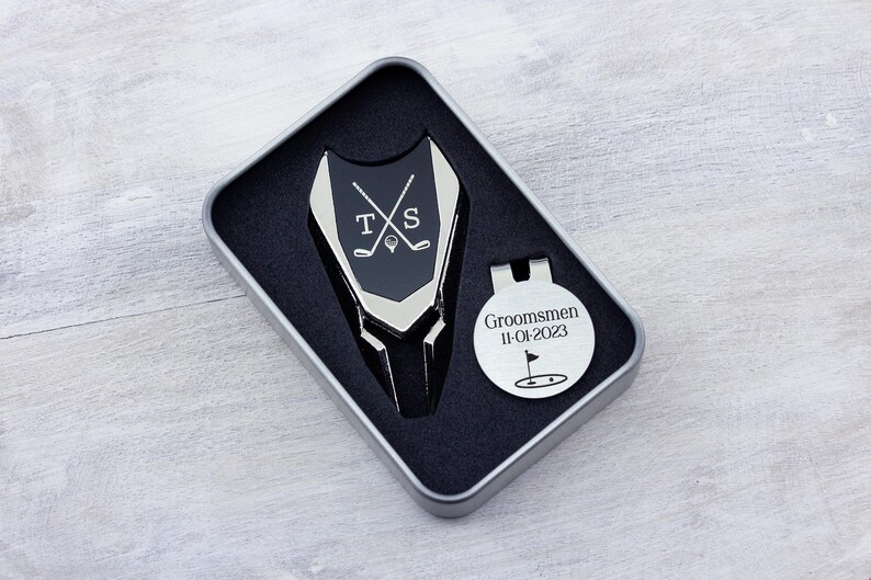 Golf ball marker and divot tool set in a gift tin box. Divot tool has marker engraved with crossed clubs with an initial on each side. The round marker comes with a hat clip and is engraved Groomsmen with the date and golf green graphic.