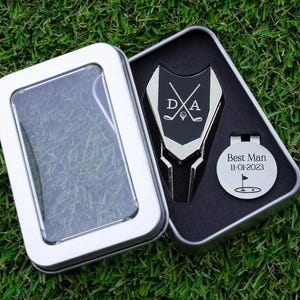 Golf ball marker and divot tool set in a gift tin box. Divot tool has marker engraved with crossed clubs with an initial on each side. The round marker comes with a hat clip and is engraved Best Man with the date and golf green graphic.