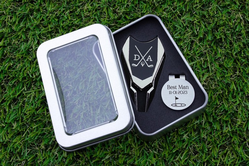 Golf ball marker and divot tool set in a gift tin box. Divot tool has marker engraved with crossed clubs with an initial on each side. The round marker comes with a hat clip and is engraved Best Man with the date and golf green graphic.
