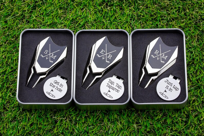 Golf ball marker and divot tool set in a gift tin box. Divot tool has marker engraved with crossed clubs with an initial on each side. The round marker comes with a hat clip and is engraved with a silly phrase and golf green graphic.