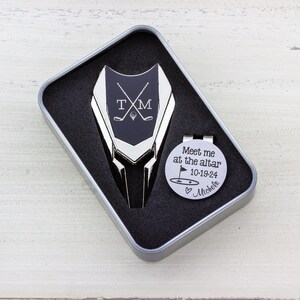 Golf ball marker and divot tool set in a gift tin box. Divot tool has marker engraved with crossed clubs with an initial on each side. The round marker comes with a hat clip and is engraved Meet me at the altar with the date, golf green graphic and a heart with name at the bottom.