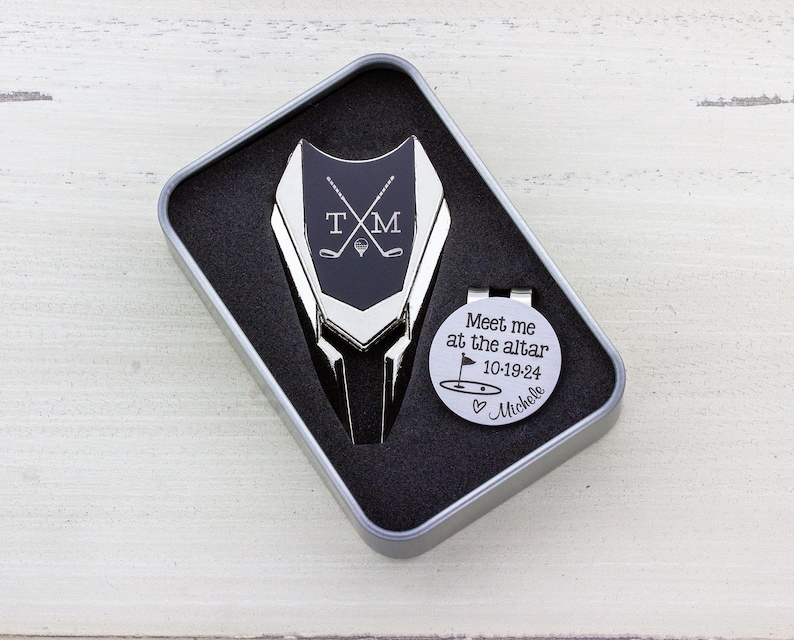 Golf ball marker and divot tool set in a gift tin box. Divot tool has marker engraved with crossed clubs with an initial on each side. The round marker comes with a hat clip and is engraved Meet me at the altar with the date, golf green graphic and a heart with name at the bottom.