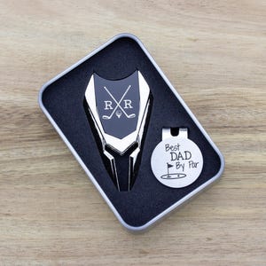 silver color metal divot tool with black metal divot tool ball marker personalized with crossed clubs with initials on each side. Silver stainless steel golf marker engraved best dad by par with golf flag graphic.