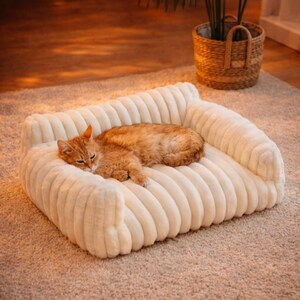 May include: A cream-colored, plush pet bed with a ribbed design. An orange tabby cat is curled up on the bed, appearing relaxed. The bed has a sofa-like shape with armrests. A woven basket with a plant is in the background.