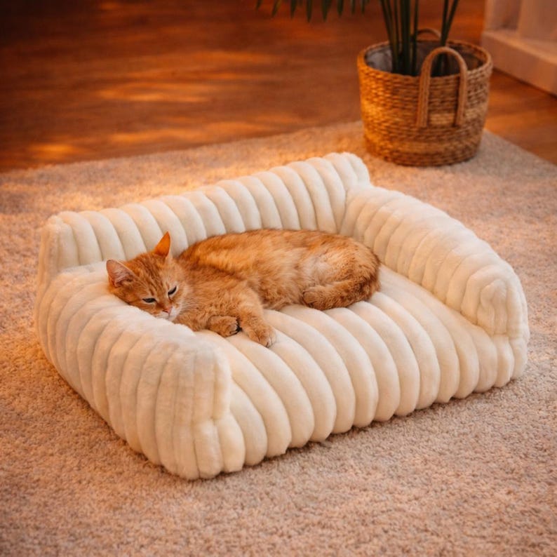 May include: A cream-colored, plush pet bed with a ribbed design. An orange tabby cat is curled up on the bed, appearing relaxed. The bed has a sofa-like shape with armrests. A woven basket with a plant is in the background.
