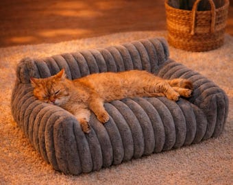 May include: A sleeping orange cat rests on a gray, plush pet sofa. The sofa has a ribbed design and is placed on a light-colored carpet. A woven basket with a plant is in the background.