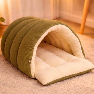 Puede incluir: Cama para mascotas verde oliva con diseño de túnel. La cama presenta un exterior de pana y un interior suave de color crema. Tiene una base rectangular y una parte superior arqueada y redondeada.