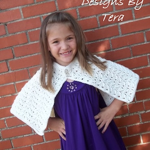 May include: A young girl wearing a white crocheted cape over a purple dress. The cape has a button fastening and is made with a delicate stitch pattern. The girl is smiling and looking at the camera. The text "Designs By Tera" is visible in the top right corner of the image.